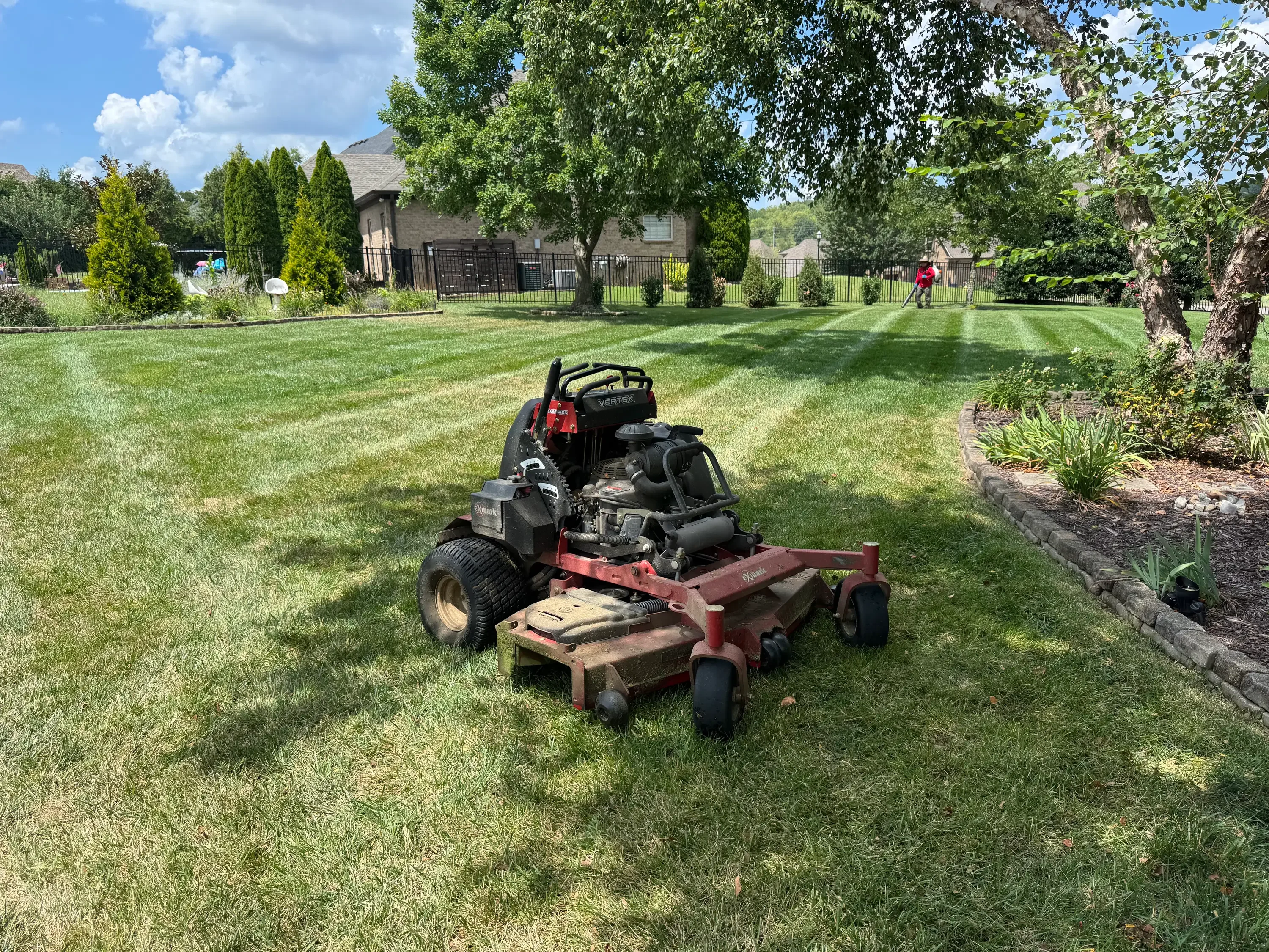 Zero-turn mower on striped residential lawn with mature trees and landscape beds